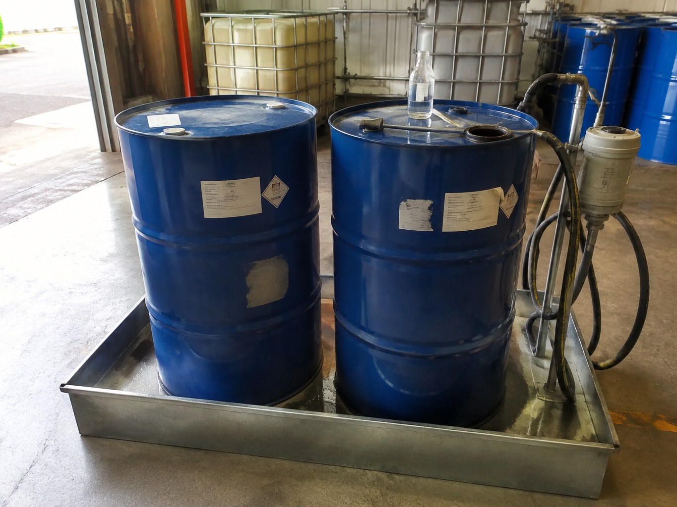 Blue solvent drums and a clear 1,2,4-trimethylbenzene sample beside warehouse transfer equipment