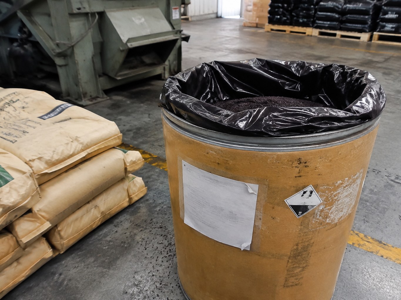 Open fibre drum of dark 6PPD granules beside rubber-compounding sacks and equipment.