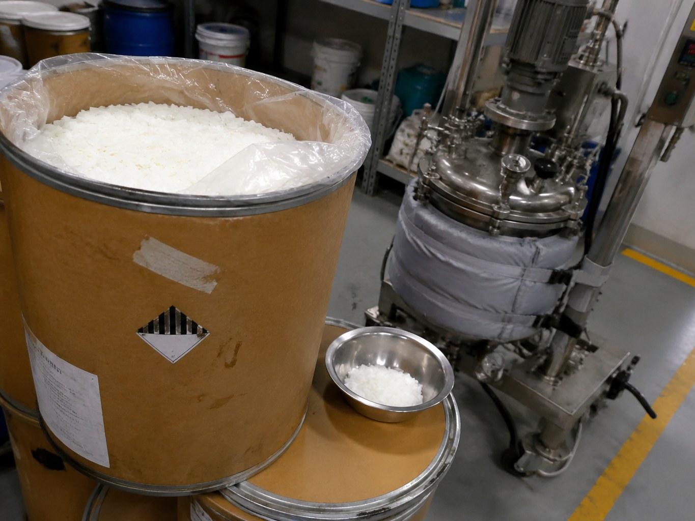 Open fibre drum of white behenic acid flakes beside oleochemical processing equipment.