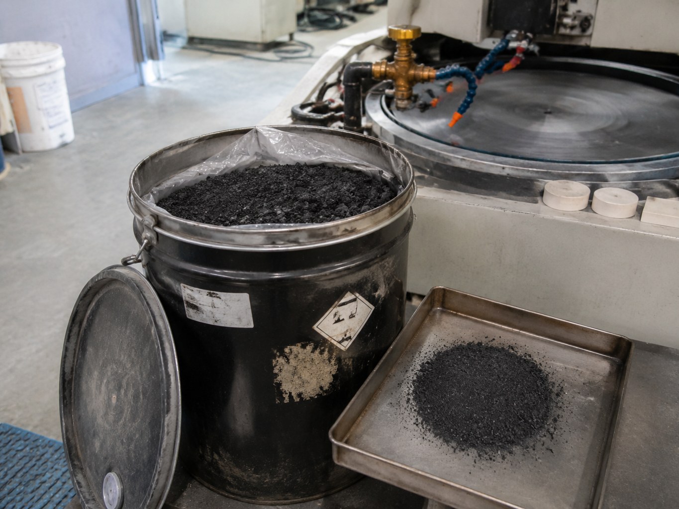 Black boron carbide powder in an open pail beside abrasive-processing equipment.