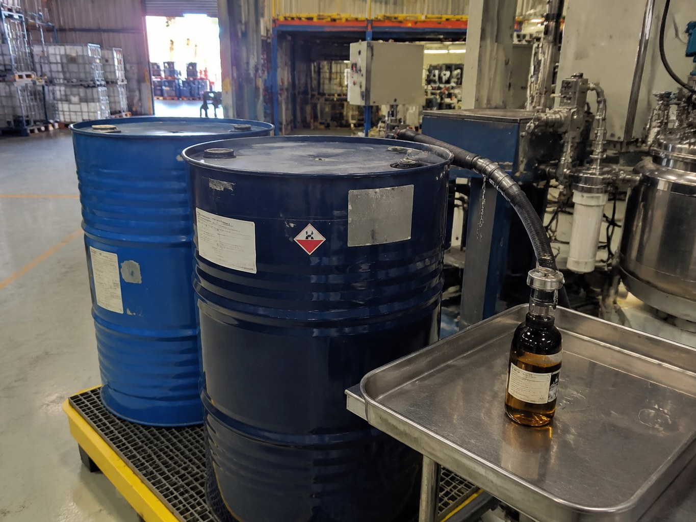 Blue butylethanolamine drums and a sample bottle beside a stainless blending vessel.