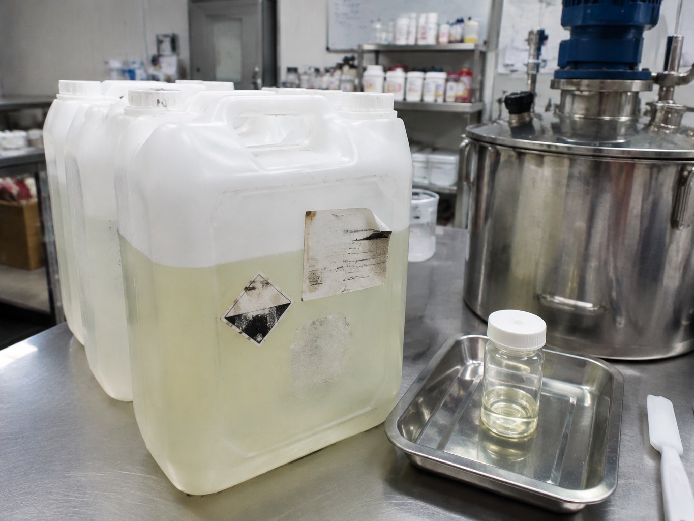 Translucent jerrycans of pale coco-caprylate/caprate beside personal-care mixing equipment.