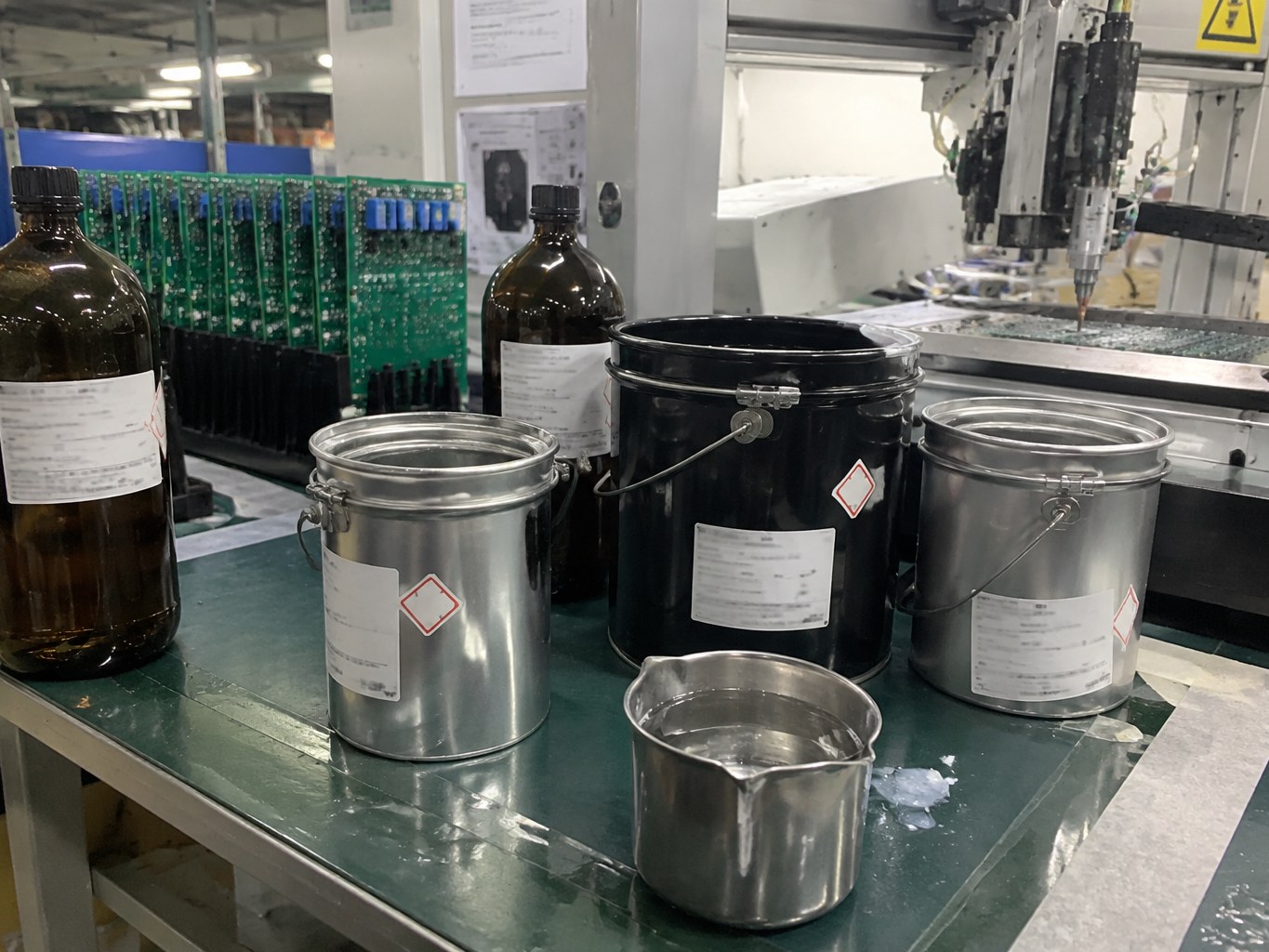 Conformal coating bottles and metal pails arranged beside electronics coating equipment.