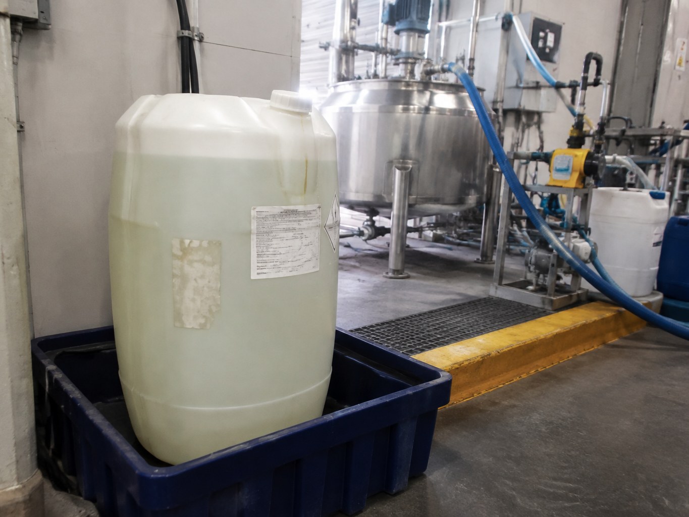 Translucent diethylethanolamine drum in containment beside stainless chemical blending equipment.