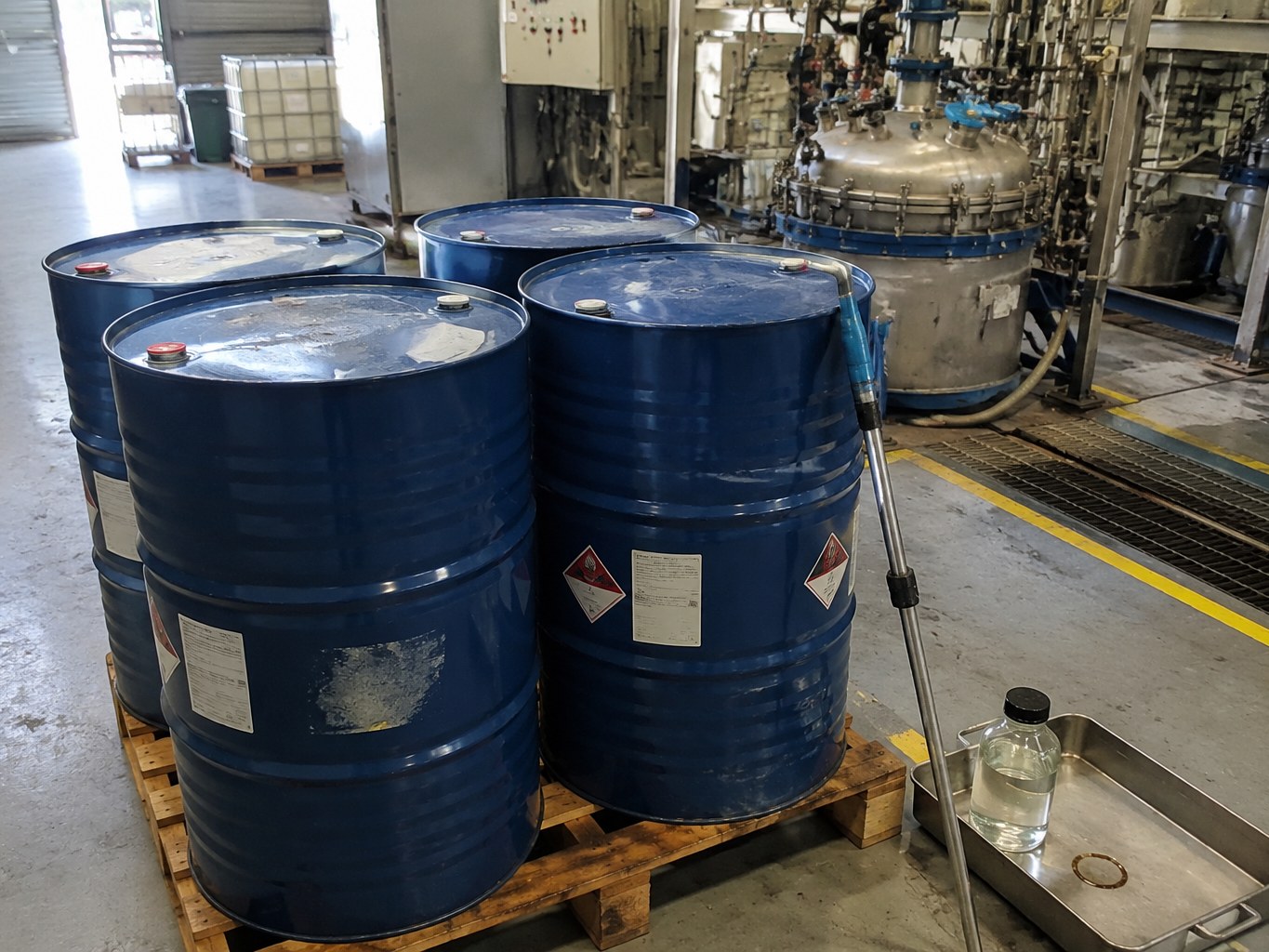 Blue drums of ethyl acetoacetate beside a reactor and stainless sampling tray.