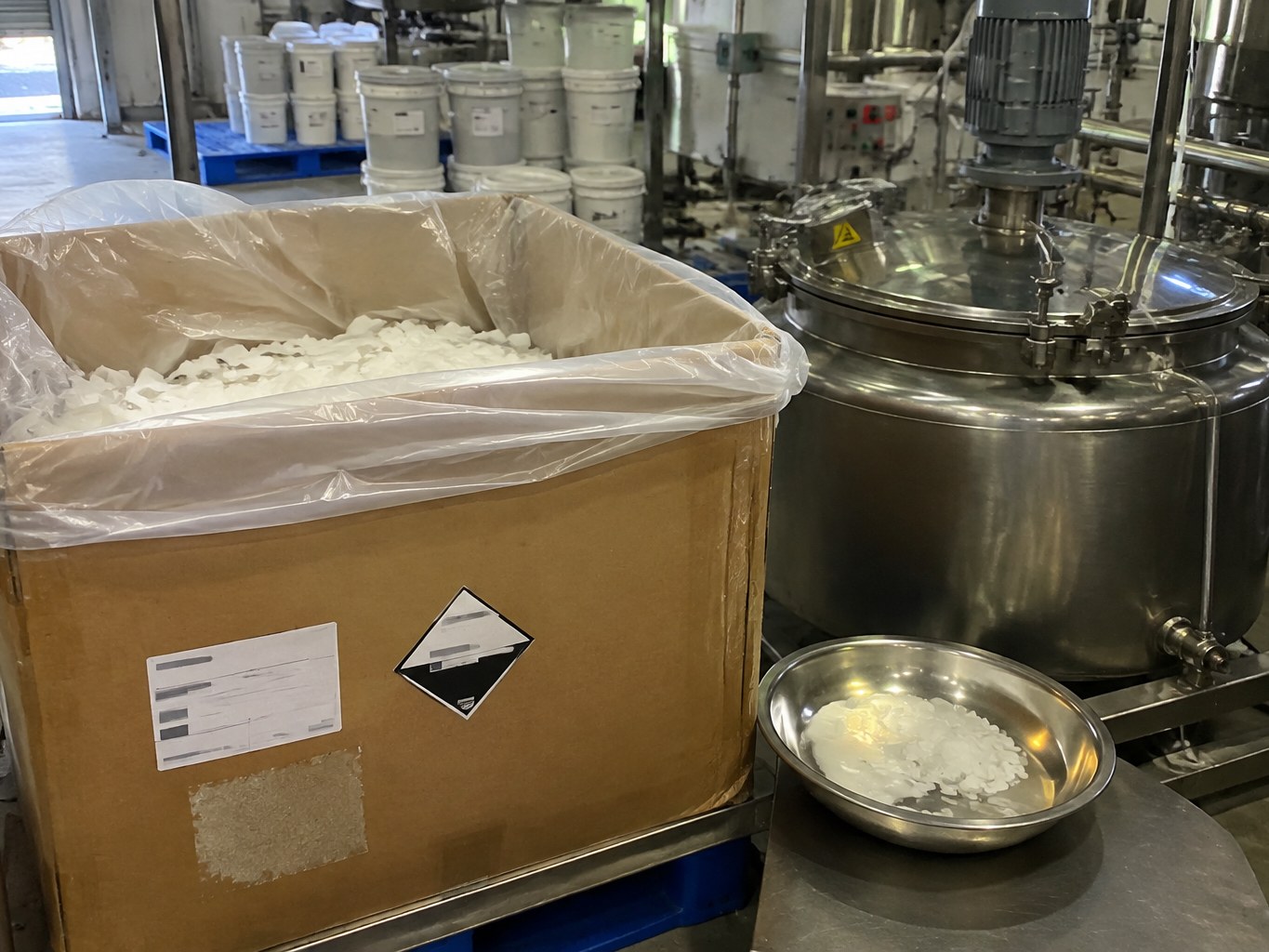 Lined carton of white glyceryl monolaurate flakes beside a formulation vessel.
