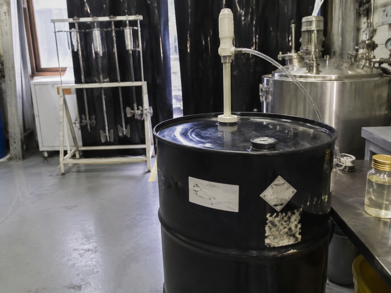 Dark drum of isobornyl acrylate beside an amber sample bottle and resin mixer