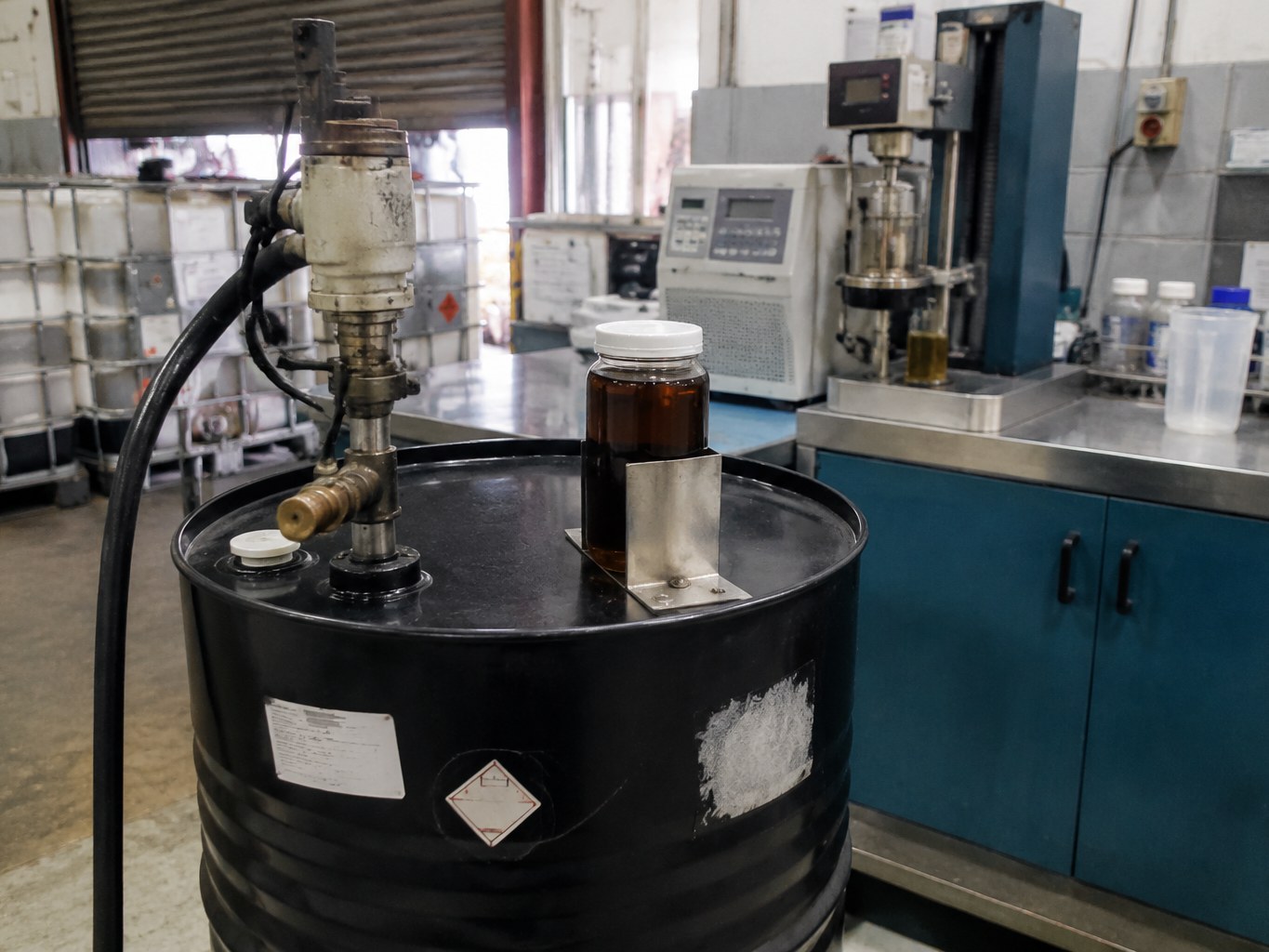 Black drum of dark molybdenum dithiocarbamate concentrate beside lubricant testing equipment.