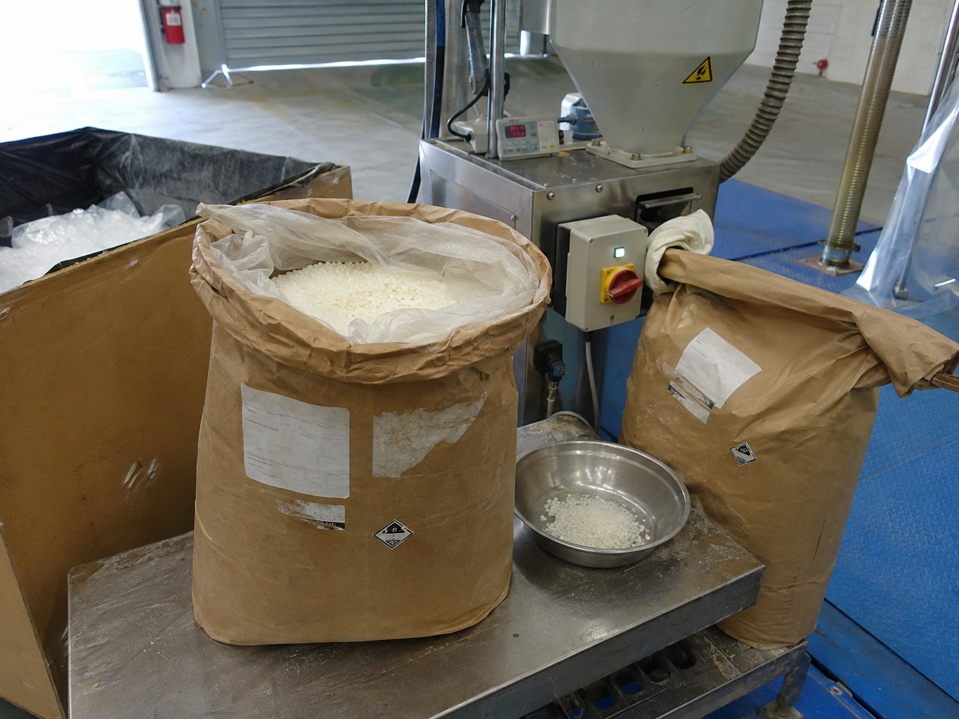 Open sack of white oleamide granules beside a plastics additive feeder