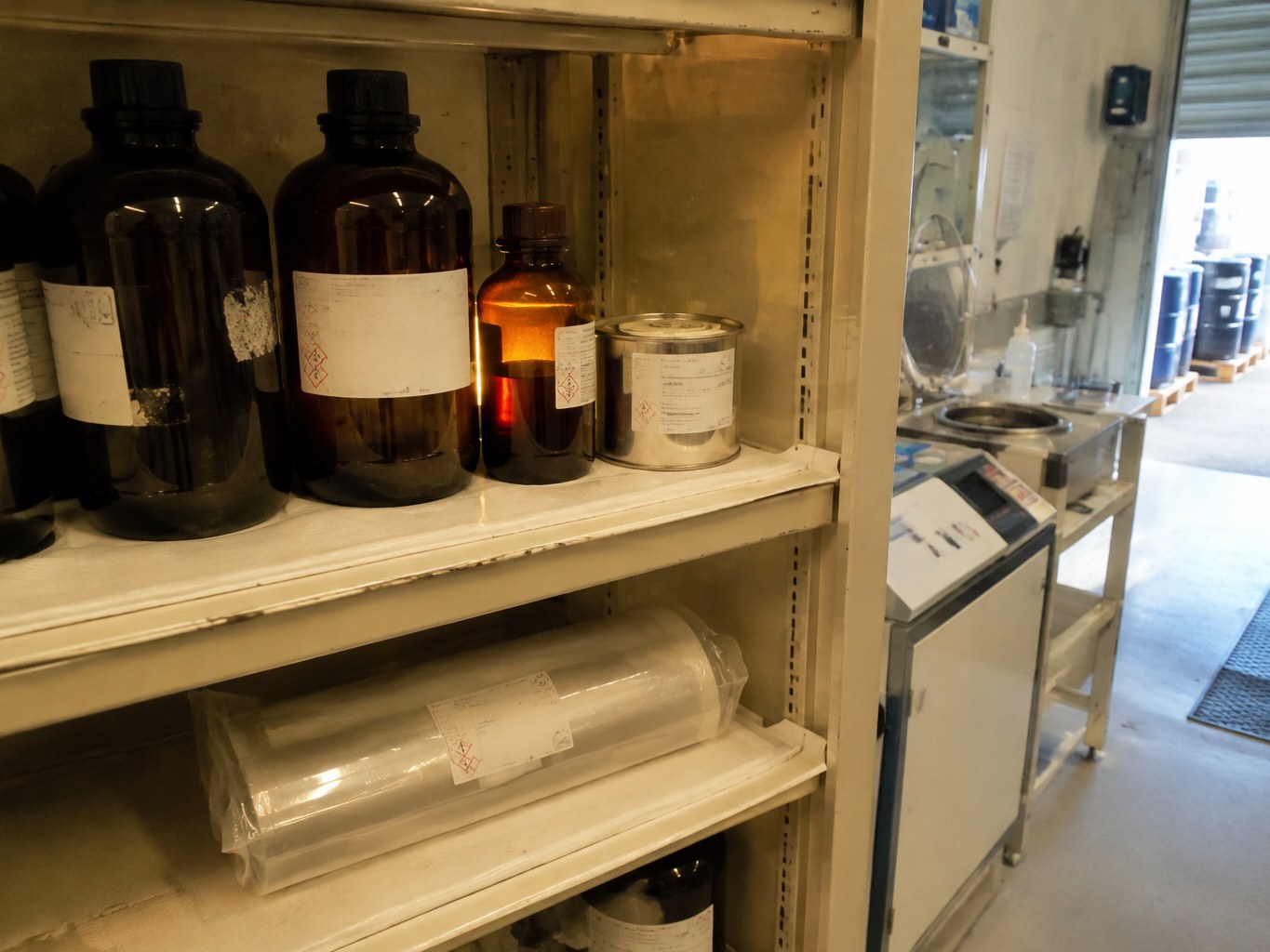 Amber photoresist bottles and protected dry-film roll in a yellow-lit electronics chemical cabinet.