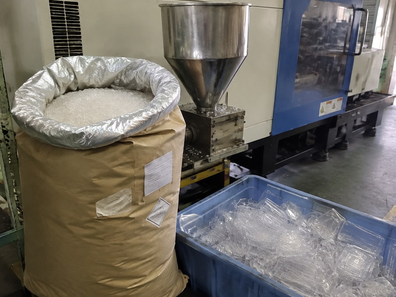 Clear SAN resin pellets in a lined sack beside an injection moulding hopper.