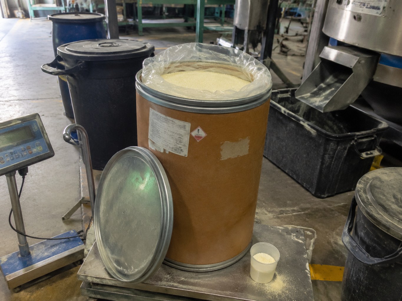Open fibre drum of pale TBBS granules beside a rubber-compounding scale.